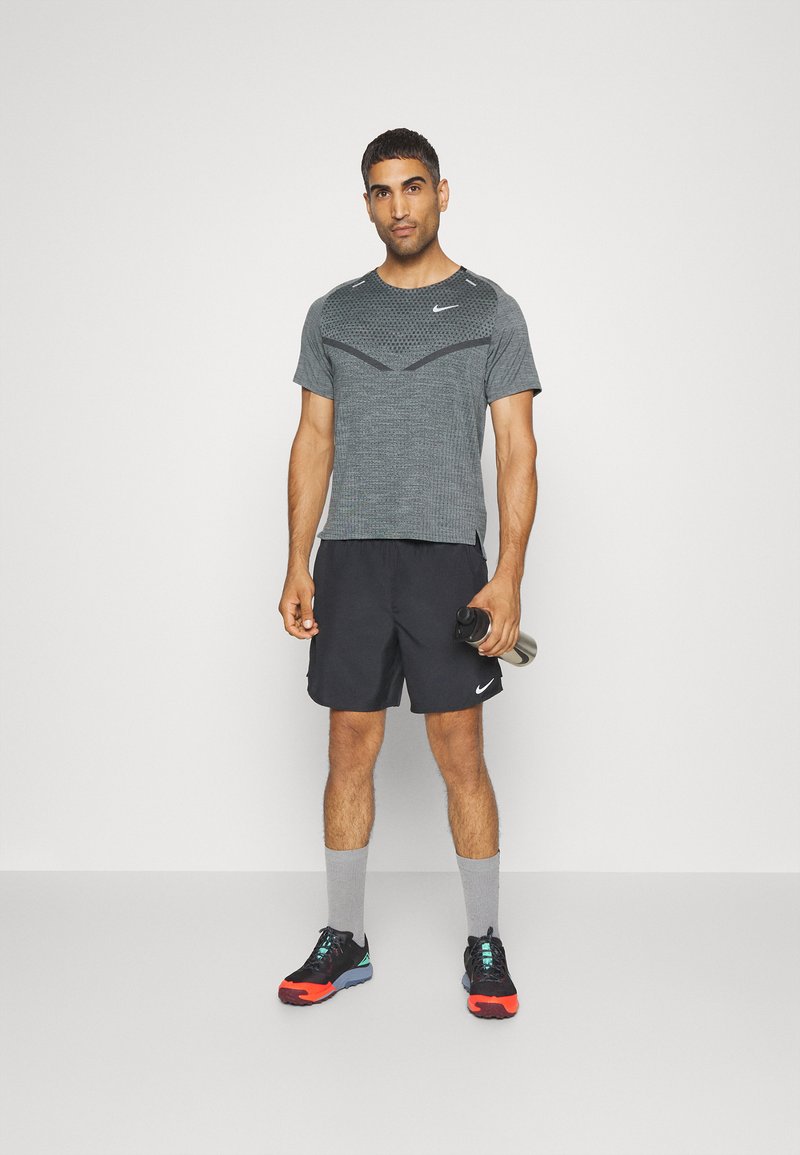 Graues Sport-T-Shirt mit Netzoberteilen, schwarzen Akzenten, kombiniert mit schwarzen Shorts. Trägt graue Socken und schwarz/orange Laufschuhe, während er eine Wasserflasche hält.