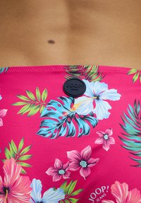 Shorts de bain roses avec des motifs floraux tropicaux, présentant des feuilles bleues et vertes, une étiquette logo noire et une texture de tissu lisse.
