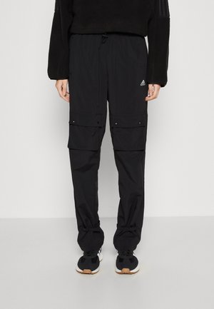 Persoon draagt zwarte Adidas cargobroek met elastische boorden en zwarte sneakers, staand tegen een effen lichte achtergrond. Bovenlichaam niet zichtbaar.