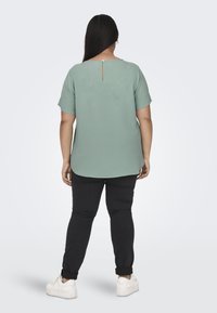 Blusa verde menta con maniche corte, scollo rotondo e dettaglio a chiave sulla schiena, abbinata a pantaloni neri e sneakers bianche.