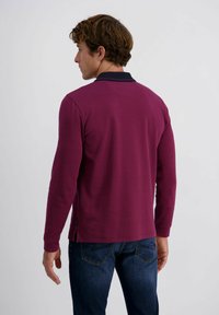 Junger Mann, der seitlich steht und ein langärmliges burgunderfarbenes Poloshirt sowie dunkelblaue Jeans trägt, vor einem schlichten hellen Hintergrund.