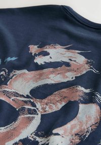 Stilisierte Drachen-Grafik in gedämpften Rosa- und Weißtönen auf dunkelblauem Stoff, die den Kopf und den verlängerten Körper des Drachens zeigt.