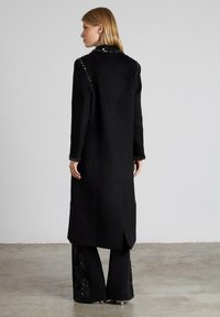 Abrigo largo negro con una tela texturizada, adornado con lentejuelas a lo largo de los hombros y los puños. Presenta un diseño de espalda partida y una silueta recta.