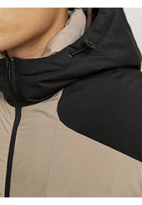 Giacca con cappuccio caratterizzata da un design a blocchi di colore nero e beige, realizzata in tessuto testurizzato con chiusura a zip e cordino regolabile nel cappuccio.