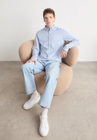 Jasnoniebieska koszula z kołnierzykiem, zestawiona z jasnymi dżinsami i białymi sneakersami, siedząca na teksturowanym beżowym krześle.