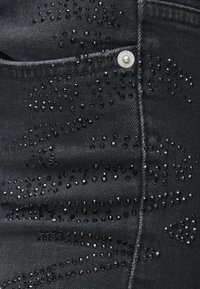 Jeans de denim preto com um padrão texturizado de pequenas contas e cristais pretos brilhantes dispersos pelo tecido. Detalhe em botão prateado visível.