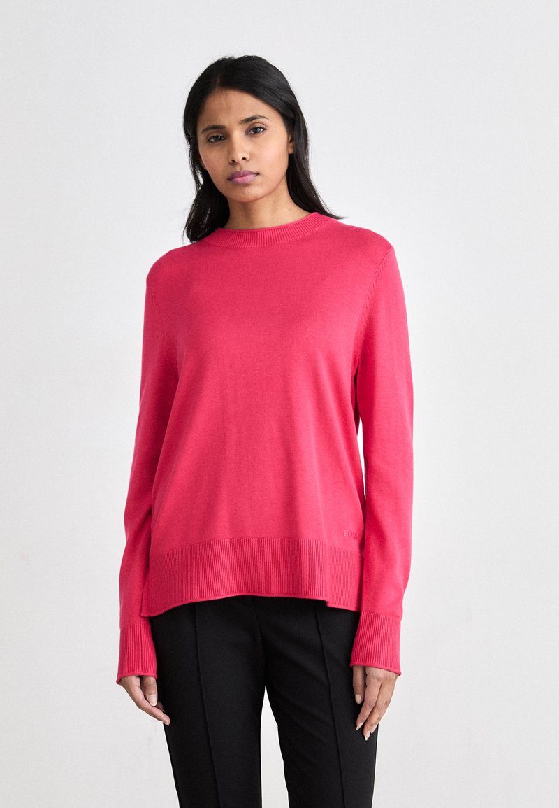 Roza pletena jopica s krojem na vratu in dolgimi rokavi, z rebrastimi detajli na robu in manšetah. V kombinaciji s črnimi hlačami.