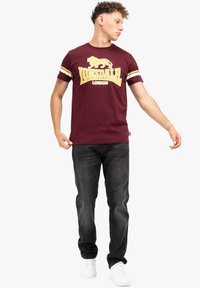 Bordo medvilnės marškinėliai su geltonomis ir baltomis juostelėmis ant rankovių, turintys liūto grafiką ir drąsiai spausdintą tekstą "Lonsdale London".