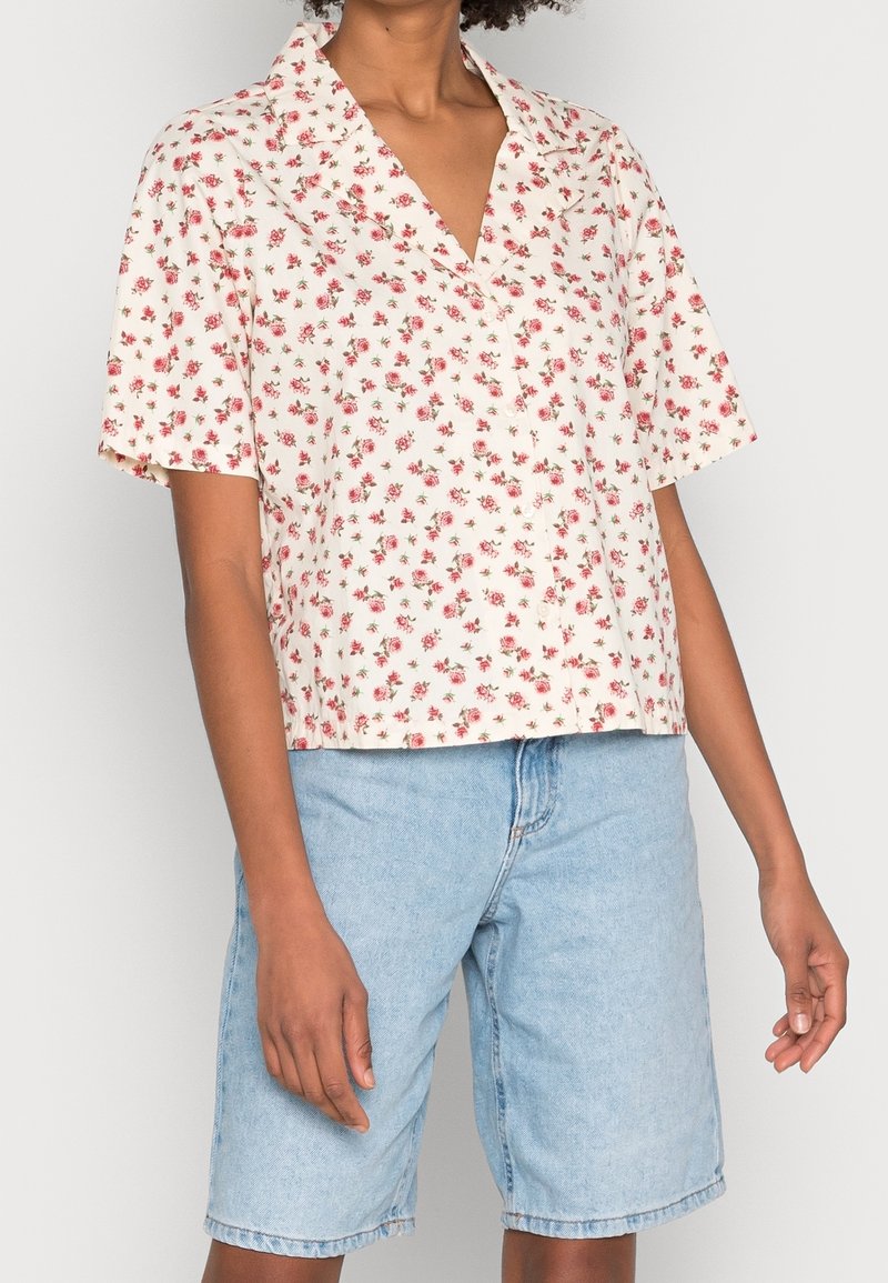 Personne portant un chemisier blanc à manches courtes avec un petit imprimé floral rouge et un short en jean bleu clair coupé aux genoux.