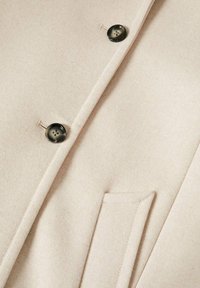 Tessuto di cappotto in misto lana crema con una texture liscia, caratterizzato da due bottoni scuri e una tasca a filetto, rifinito con cuciture precise.