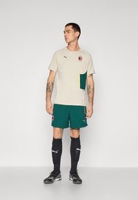 Beige atletisk skjorte med grønne og gule detaljer, parret med grønne shorts med et logo, sorte sokker og sorte sportssko.