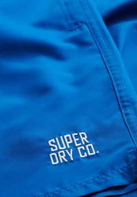 Tessuto blu con una texture liscia, caratterizzato da un logo ricamato bianco "SUPER DRY CO." e dettagli di cucitura ben visibili.