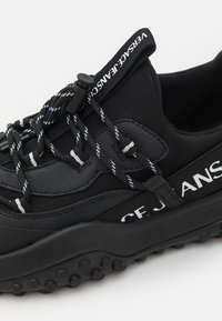 Baskets noires avec semelle épaisse, lacets à motifs, languette de tirage et marquage "Versace Jeans" sur le côté et la languette.