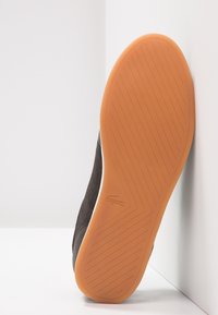 Svart mockaskosula med en orange gummisula. Har horisontella räfflor för grepp och en präglad logotyp på sidan.