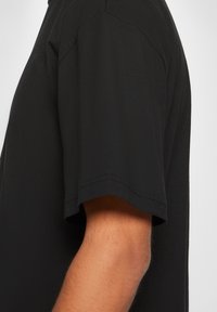 Boční pohled na ruku osoby, která má na sobě jednoduché černé tričko s krátkým rukávem proti bílému pozadí.