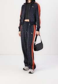 Navy patterned tracksuit with orange side stripes, zip-up jacket, and drawstring pants. Black handbag worn on arm; light sneakers.