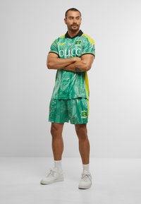 Grøn mønstrede polo- og shorts-sæt med gule accenter, sort krave og logo-detalje på shortsene. Bæres med hvide sneakers.