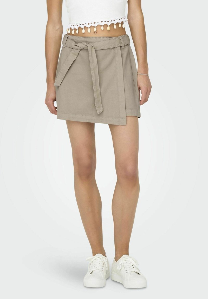 Beiger Wickelrock mit Gürtel, aus Baumwollstoff gefertigt. Verfügt über eine glatte Textur und einen geraden Schnitt, kombiniert mit weißen Turnschuhen.