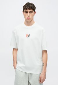 Λευκό βαμβακερό t-shirt με άνετη εφαρμογή, που διαθέτει ένα μικρό πολύχρωμο γεωμετρικό λογότυπο στη μπροστινή πλευρά. Μαλακή υφή, κοντά μανίκια.