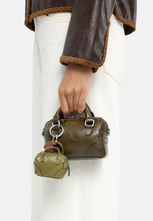 Ręka trzymająca małą torebkę ze skóry w oliwkowym kolorze z dopasowanym miniaturowym breloczkiem do kluczy, ubrana w skórzaną kurtkę z brązowym obszyciem i białe spodnie.