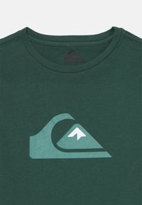 T-shirt in cotone verde con una grafica bianca e verde chiaro raffigurante un'onda e una montagna. Collo rotondo con una texture morbida.