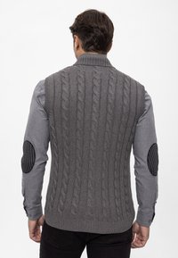 Grijze gebreide vest met kabelpatroon, geribbelde kraag en textuurelle elleboogpatches. Draag over een lange mouw grijs shirt. Subtiel contrast in materialen.