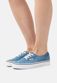 Baskets en toile bleue à surface texturée, avec des lacets blancs et une semelle noire, présentant un design classique de chaussure de skate.