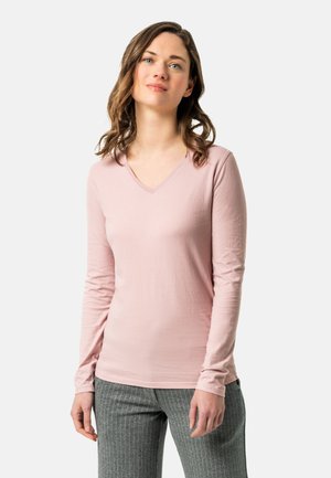 Langärmliges, V-Ausschnitt T-Shirt aus zartem rosa Stoff mit figurbetontem Schnitt und glatter Textur, kombiniert mit grauen Stoffhosen mit Nadelstreifen.