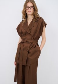 Gilet doppio petto marrone con maniche corte, cintura e due tasche frontali. Tessuto liscio e vestibilità sartoriale, abbinato a pantaloni coordinati.
