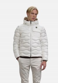 Giacca a vento bianca con un design trapuntato testurizzato, cerniera frontale, cappuccio e tasca con zip. Abbinata a pantaloni di colore chiaro.