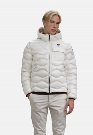 Giacca a vento bianca con un design trapuntato testurizzato, cerniera frontale, cappuccio e tasca con zip. Abbinata a pantaloni di colore chiaro.