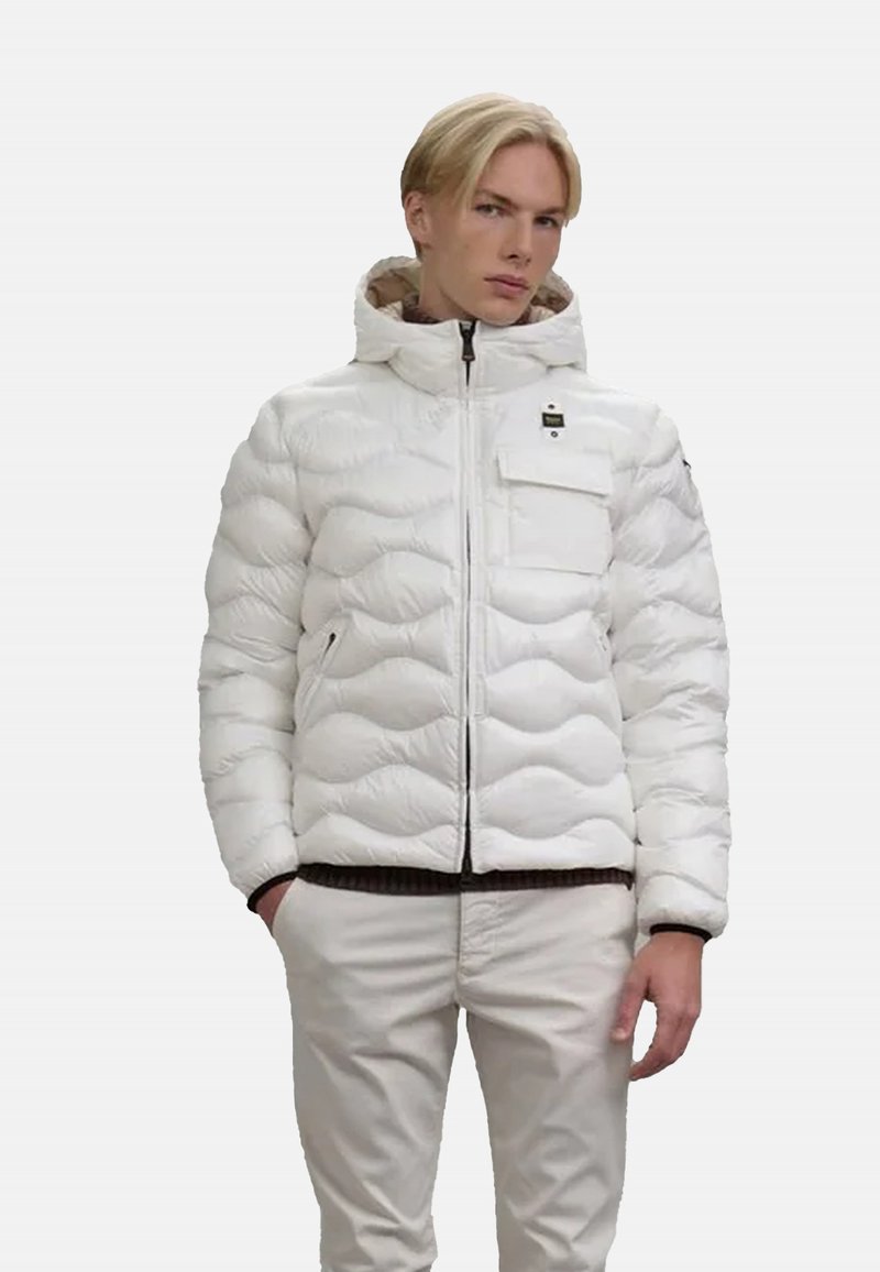 Giacca a vento bianca con un design trapuntato testurizzato, cerniera frontale, cappuccio e tasca con zip. Abbinata a pantaloni di colore chiaro.