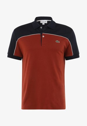 Rode polo shirt met marineblauwe accenten, korte mouwen en kraag. Bevat witte piping en een groene Lacoste-logo op de linkerborst.