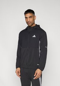 Μαύρο μπουφάν με φερμουάρ και κουκούλα, με γκρι πλαϊνά σχέδια και λευκό λογότυπο στο στήθος. Κατασκευασμένο από ελαφρύ, λείο ύφασμα, με ελαστικές μανσέτες.