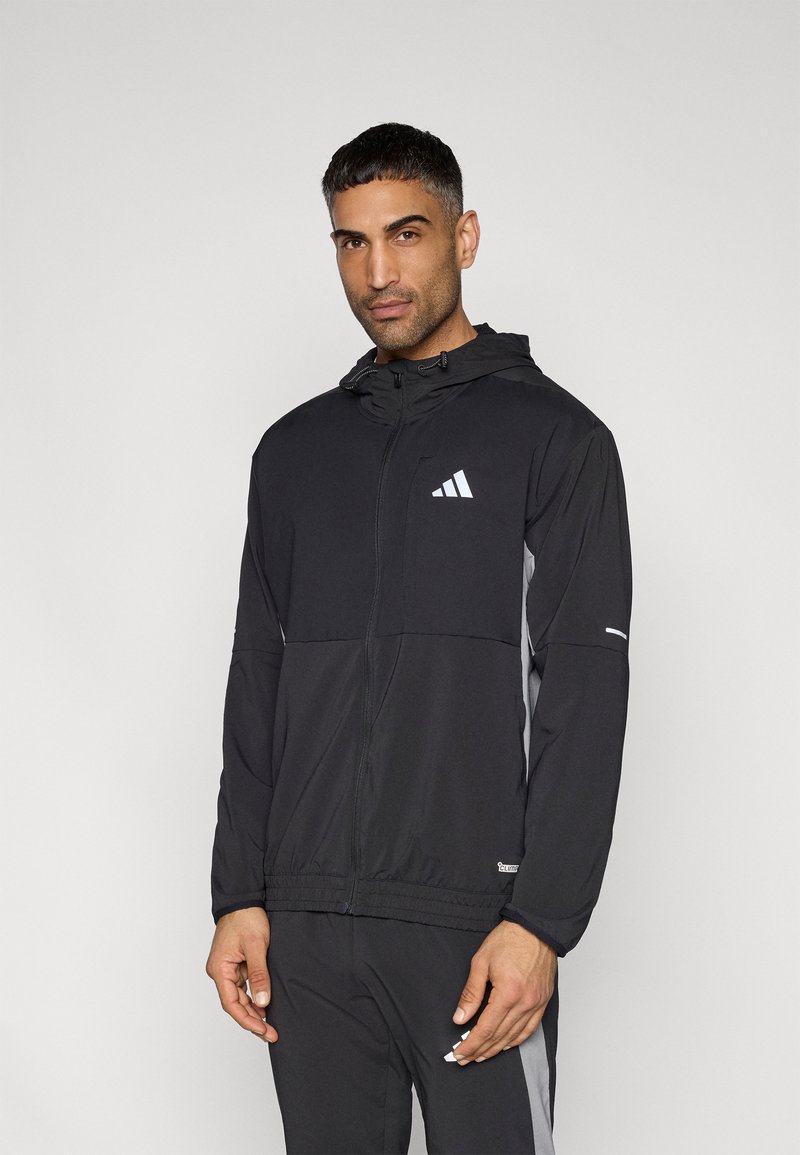 Μαύρο μπουφάν με φερμουάρ και κουκούλα, με γκρι πλαϊνά σχέδια και λευκό λογότυπο στο στήθος. Κατασκευασμένο από ελαφρύ, λείο ύφασμ α, με ελαστικές μανσέτες.