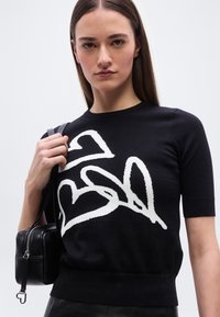 Γυναίκα που φοράει μαύρο μπλουζάκι με κοντά μανίκια και μεγάλα άσπρα αφηρημένα σχέδια καρδιάς, κρατώντας μια μαύρη δερμάτινη τσάντα στον ώμο της.