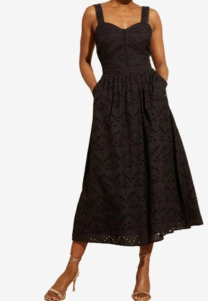 Donna che indossa un abito midi nero senza maniche con ricamo ad occhio, corpo attillato e tasche, abbinato a tacchi beige con cinturini.