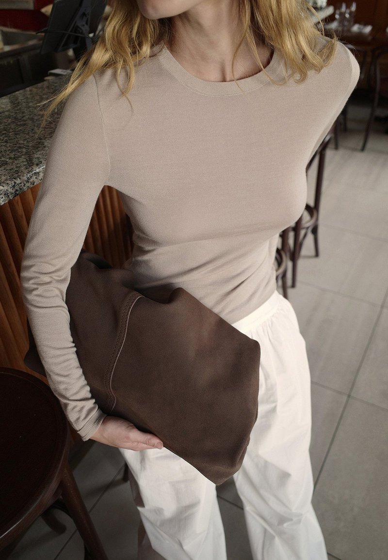 Beige pitkähihaine paita, valkoiset housut, pitää isoa ruskeaa mokkakirjettä. Suojaava pinta, yksinkertainen muotoilu, jossa näkyvät ompeluseikat.
