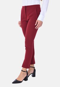 Pantaloni tailored bordeaux con una vestibilità slim e tasche laterali; presentano una chiusura lampo frontale e una vita piatta, abbinati a scarpe nere con tacco a blocco.