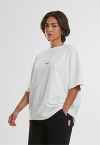 T-shirt en coton oversized blanc à manches courtes, avec un texte rouge brodé sur la poitrine et une coupe décontractée. Associé à un pantalon noir texturé.