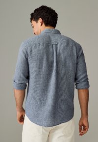 Muška plava teksturirana košulja na kopčanje s podvrnutim rukavima, s naborom na leđima i zaobljenim rubom, izrađena od laganog tkanja.