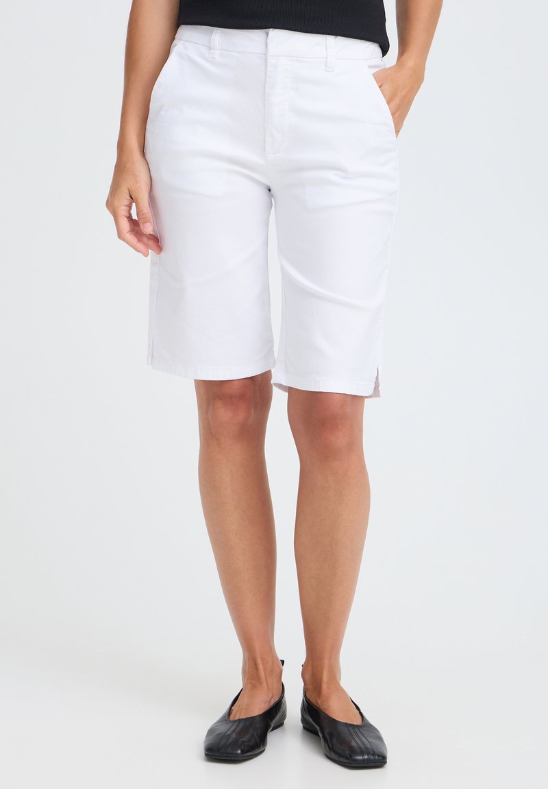 Femme portant un short blanc au genou, un haut noir et des chaussures plates noires, debout avec une main dans la poche devant un fond uni.