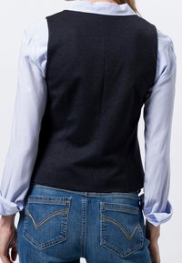 Ciemnoszara dopasowana kamizelka z gładkiego materiału, noszona na jasnoniebieskiej koszuli w paski, zestawiona z niebieskimi dżinsami. Widoczne detale przeszyć.