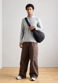 Γκρι μακρυμάνικο πουκάμισο με φερμουάρ, καφέ φαρδιά παντελόνια, μαύρη τσάντα ώμου και λευκά αθλητικά παπούτσια· στέκεται σε ξύλινο πάτωμα σε ουδέτερο χώρο.