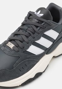 Grå atletisk sneakers som har en blandning av mocka och mesh-material, tre vita ränder, vadderad krage och en kraftig kontrasterande sula.