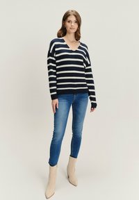 Sweter w niebiesko-białe paski z dekoltem w serek, wykonany z miękkiego materiału; zestawiony z dopasowanymi niebieskimi dżinsami i beżowymi botkami typu ankle.
