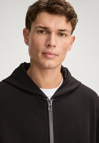 Zwarte zip-up hoodie met een textuurstof, een grote capuchon en een metalen ritssluiting. Draag over een witte shirt.