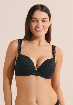 Smiling woman wearing a black lace bra with thin straps standing against a plain beige background.