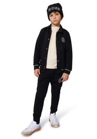 Sweat-shirt noir et joggings avec accents de logo, t-shirt crème, et bonnet noir. Baskets blanches et grises avec des bandes Velcro et semelles texturées.