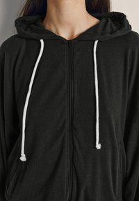 Detailný záber osoby v čiernej mikine na zips s bielymi šnúrkami a mierne voľným strihom.